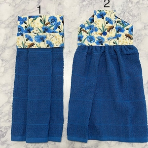 May include: Two blue kitchen towels with a floral and bee pattern at the top. The towels are a solid blue color with a textured weave. The top section features a cream-colored fabric with blue flowers and bee illustrations.