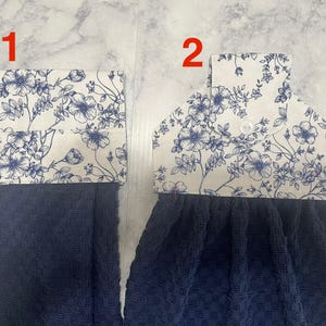May include: Two navy blue kitchen towels with a floral design. The top portion of each towel features a white fabric with blue floral patterns. One towel has a simple folded top, while the other has a button closure.