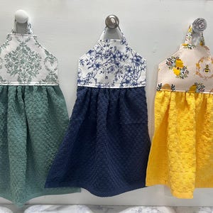 May include: Three hanging kitchen towels with fabric tops and textured bottoms. The first has a green damask top and green towel, the second a blue floral top and navy towel, and the third a bee-patterned top with a bright yellow towel.