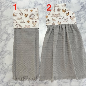 May include: Two gray kitchen towels with a decorative top featuring a chicken and coop pattern. The towels have a textured design and fringe at the bottom. One towel is folded over, and the other is hanging.