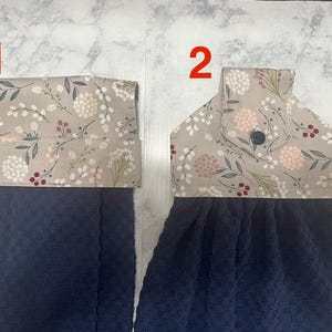 May include: Two navy blue kitchen towels with floral patterned tops. The tops feature a beige background with pink, white, and burgundy floral designs. One towel has a folded top, and the other has a button closure.