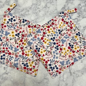 May include: Two white cotton pot holders with a floral pattern in shades of red, blue, yellow, and pink. The pot holders have a loop handle.