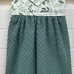 May include: A hanging kitchen towel featuring a dark teal, textured waffle-weave fabric attached to a white fabric topper with a green leaf and pink dot pattern. The topper includes a single white button for securing the towel to a handle.