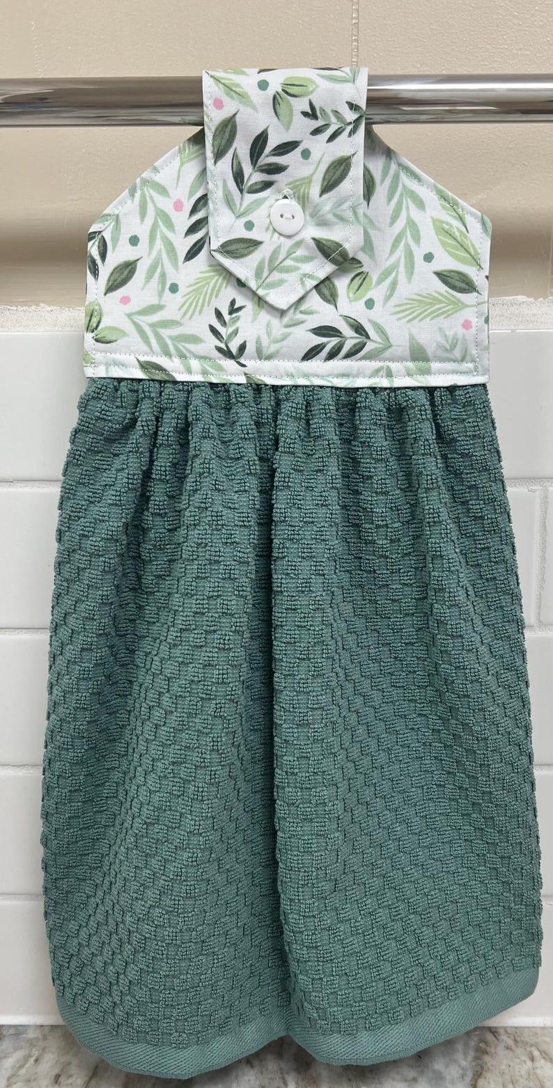 May include: A hanging kitchen towel featuring a dark teal, textured waffle-weave fabric attached to a white fabric topper with a green leaf and pink dot pattern. The topper includes a single white button for securing the towel to a handle.