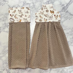 May include: Two hanging kitchen towels with a beige waffle-weave texture. The top sections feature a cream-colored fabric printed with various chickens, roosters, and farm structures. One towel includes a button closure tab at the top center.