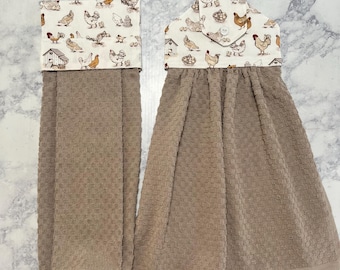 May include: Two hanging kitchen towels with a beige waffle-weave texture. The top sections feature a cream-colored fabric printed with various chickens, roosters, and farm structures. One towel includes a button closure tab at the top center.