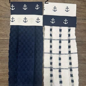 May include: Two kitchen towels with anchor print fabric bands. The towel on the left is dark blue with a waffle texture. The towel on the right is white with black and white checkered pattern.