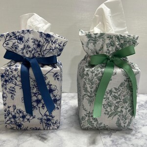 May include: Two square fabric tissue box covers with white backgrounds. The left cover features a blue floral pattern and a dark blue ribbon bow. The right cover features a green floral pattern and a light green ribbon bow. Both have tissues.