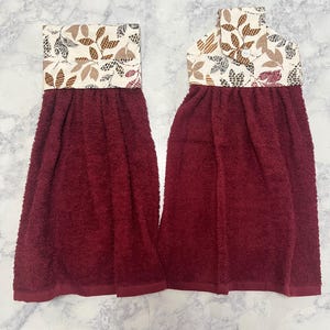 May include: Two hanging kitchen towels featuring deep burgundy terry cloth fabric. The top sections are made of cream-colored fabric with a brown and dark red leaf pattern. One towel has a folded top with a white button closure for hanging.