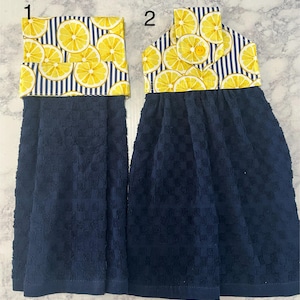May include: Two blue kitchen towels with lemon print fabric accents. The towels are hanging on a white surface. The top towel has a button and the bottom towel has a striped fabric accent.