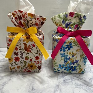 May include: Two fabric tissue box covers with white tissues protruding from the top. The left cover features a squirrel and autumn floral print with a yellow ribbon. The right cover features a blue and pink floral print with a hot pink ribbon.
