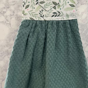 May include: A hanging kitchen towel featuring a textured, solid teal-colored fabric base attached to a white fabric top panel. The top panel displays a botanical pattern of green leaves and small pink dots, secured by a single white button closure.