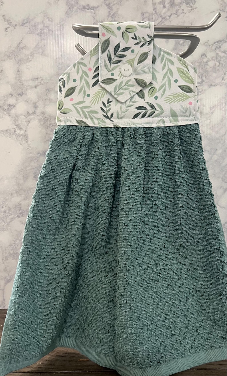May include: A hanging kitchen towel featuring a textured, solid teal-colored fabric base attached to a white fabric top panel. The top panel displays a botanical pattern of green leaves and small pink dots, secured by a single white button closure.