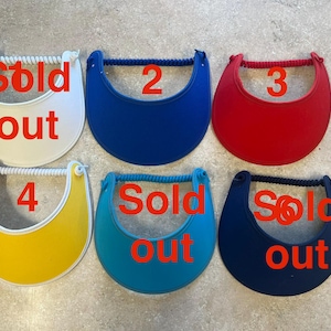 May include: Six sun visors arranged in two rows, featuring a curved brim and a coiled headband. The colors include white, blue, red, yellow, teal, and navy. The white, teal, and navy visors are marked with the red text Sold out.