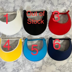 May include: Six visors in various colors: white, black, red, yellow, blue, and navy. Each visor has a curved brim and a headband. The black visor has the text "Out of Stock".