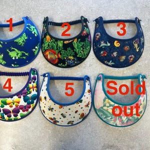 May include: Six sun visors with elastic back straps, numbered one through five and one marked Sold out. Designs include green frogs on blue, tropical fish on black, sea creatures on dark blue, colorful butterflies, bees on white, and floral.