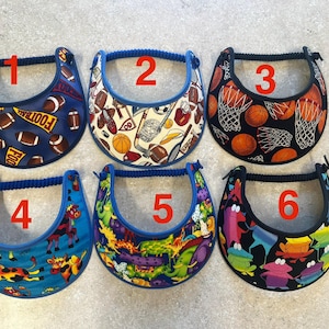 May include: Six visors with blue rope headbands. Each visor has a different pattern: footballs, sports equipment, basketballs, cartoon cows, colorful dinosaurs, and cartoon characters. The visors are designed to shield the eyes from the sun.