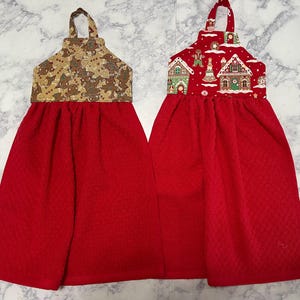 May include: Two hanging kitchen towels with red fabric bottoms. The left towel features a brown gingerbread man print on the top section. The right towel features a red top section with a gingerbread house and holiday cookie pattern.