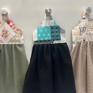 May include: Three hanging kitchen towels with fabric tops. The left has a floral print and sage green towel. The center has a teal and black geometric print with a black towel. The right has a coffee-themed print with a beige towel.