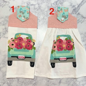 May include: Two hanging kitchen towels. Each towel features a teal truck filled with pink and red flowers. The word "BLOOM" is printed on the truck. The top of each towel is pink with white polka dots and a butterfly print.