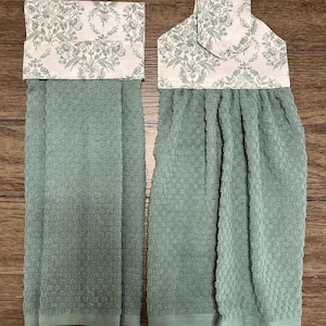 May include: Two sage green kitchen towels featuring a textured waffle weave pattern. Each towel is topped with a fabric header displaying a light cream background with a repeating sage green floral and leaf damask print design.