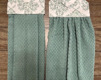May include: Two sage green kitchen towels featuring a textured waffle weave pattern. Each towel is topped with a fabric header displaying a light cream background with a repeating sage green floral and leaf damask print design.