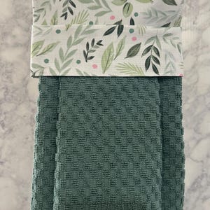 May include: A hanging kitchen towel featuring a dark green waffle-textured fabric base and a top fabric panel decorated with a pattern of green leaves and small pink dots. The towel is folded over a silver metal bar for display and storage purposes.