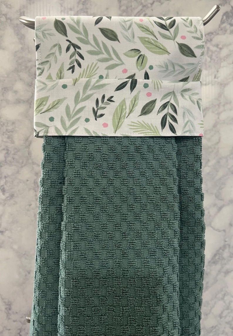 May include: A hanging kitchen towel featuring a dark green waffle-textured fabric base and a top fabric panel decorated with a pattern of green leaves and small pink dots. The towel is folded over a silver metal bar for display and storage purposes.