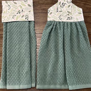 May include: Two sage green waffle-weave kitchen towels featuring a top fabric panel with a white background and a botanical print of green leaves and small pink dots. The right towel includes a fabric tab with a button for hanging purposes.