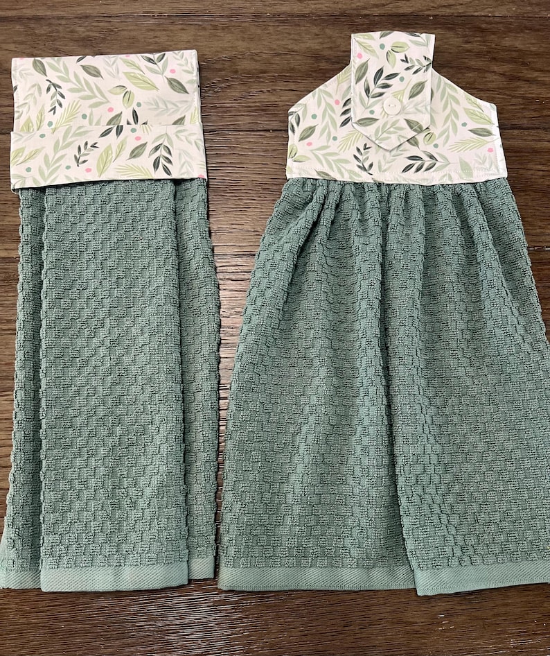 May include: Two sage green waffle-weave kitchen towels featuring a top fabric panel with a white background and a botanical print of green leaves and small pink dots. The right towel includes a fabric tab with a button for hanging purposes.