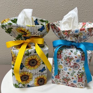 May include: Two fabric tissue box covers. The left cover features a sunflower and blue butterfly print with a yellow ribbon. The right cover features a bicycle and floral print with a bright blue ribbon. Both covers hold white facial tissues.