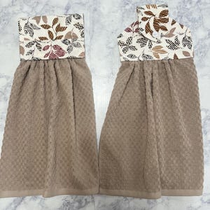 May include: Two hanging kitchen towels with a textured tan waffle-weave fabric. The top sections feature a cream-colored fabric with a printed pattern of brown, black, and maroon leaves. One towel includes a small white button closure at the top.