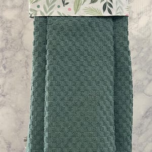 May include: A hanging kitchen towel featuring a solid sage green waffle-textured fabric on the bottom half and a white fabric top section decorated with a pattern of green leaves and small pink dots, folded over a silver metal towel bar.