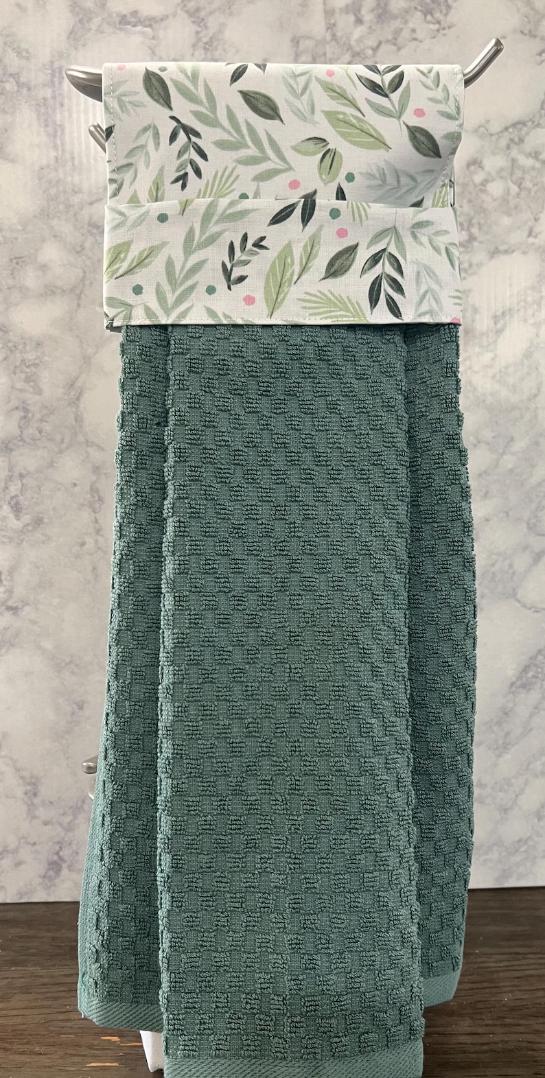 May include: A hanging kitchen towel featuring a solid sage green waffle-textured fabric on the bottom half and a white fabric top section decorated with a pattern of green leaves and small pink dots, folded over a silver metal towel bar.