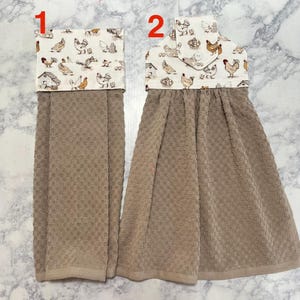 May include: Two hanging kitchen towels. The towels have a tan, textured body and a white top with a chicken and coop pattern. One towel has a straight top, and the other has a folded top with a button.