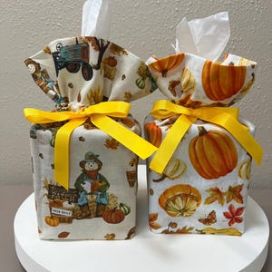 May include: Two fabric tissue box covers featuring autumn themes. The left cover displays a tractor, scarecrow, and text reading Pumpkin Patch Open Daily. The right cover features orange pumpkins and fall leaves. Both are tied with bright yellow ribbons.