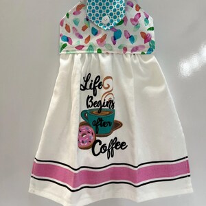 May include: White hanging kitchen towel with a colorful floral pattern and a turquoise polka dot top. The towel features the text "Life Begins after Coffee" with a coffee cup and donut graphic, and a pink stripe.