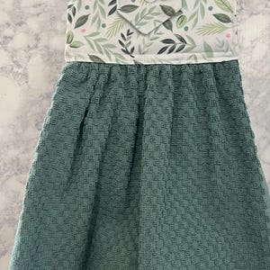 May include: A hanging kitchen towel featuring a solid dark teal textured fabric bottom and a white top panel decorated with a pattern of green leaves and small pink dots. The top section includes a white button closure for hanging on a rack.