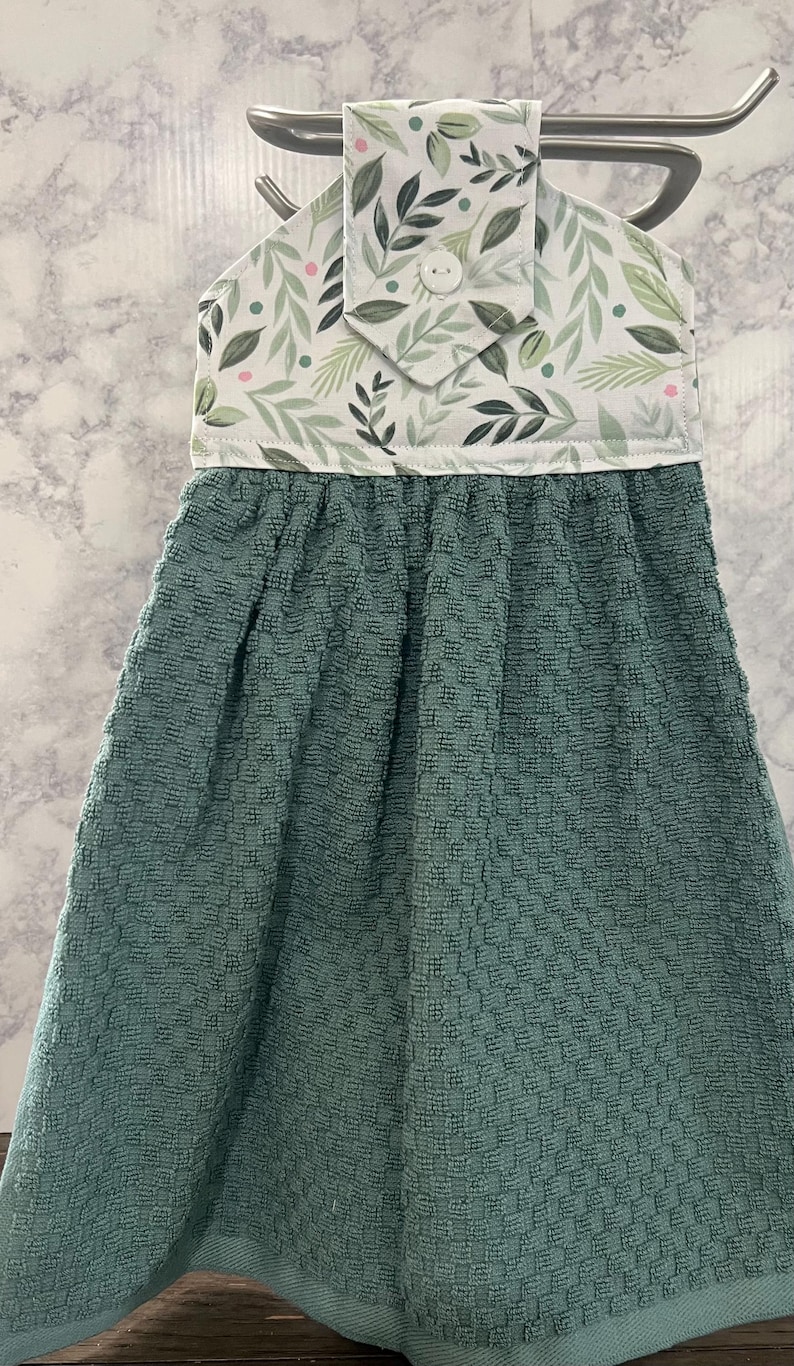 May include: A hanging kitchen towel featuring a solid dark teal textured fabric bottom and a white top panel decorated with a pattern of green leaves and small pink dots. The top section includes a white button closure for hanging on a rack.