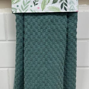 May include: A hanging kitchen towel with a textured, dark teal waffle-weave fabric. The top section features a white fabric band decorated with a botanical pattern of green leaves and small pink dots, designed to fold over a towel bar.