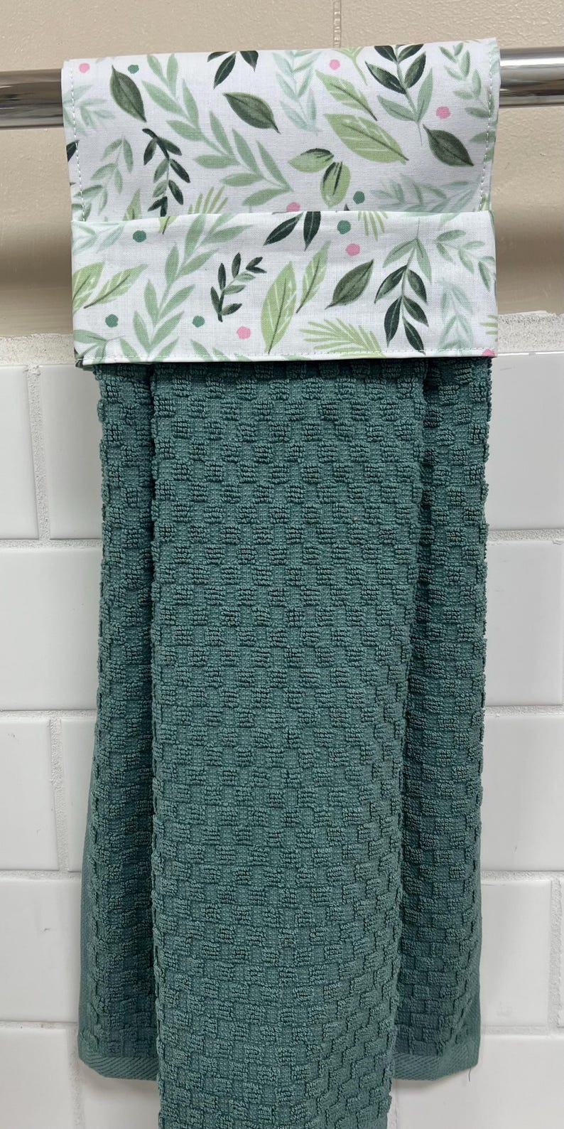 May include: A hanging kitchen towel with a textured, dark teal waffle-weave fabric. The top section features a white fabric band decorated with a botanical pattern of green leaves and small pink dots, designed to fold over a towel bar.