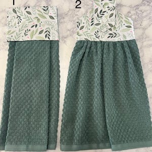 May include: Two hanging kitchen towels with a textured, dark sage green waffle weave fabric. The top sections feature a white fabric with a botanical print of green leaves and small pink dots. Towel 2 includes a white button closure at the top.