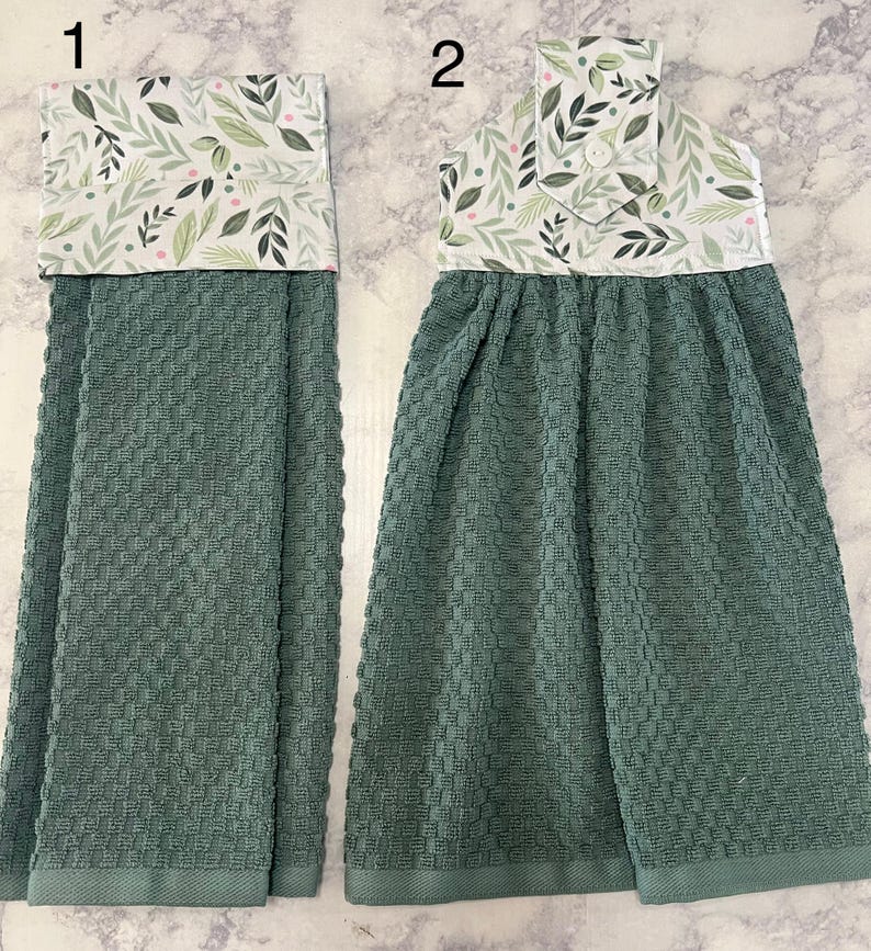 May include: Two hanging kitchen towels with a textured, dark sage green waffle weave fabric. The top sections feature a white fabric with a botanical print of green leaves and small pink dots. Towel 2 includes a white button closure at the top.
