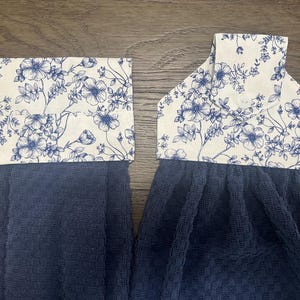May include: Two hanging kitchen towels featuring a dark navy blue waffle-textured fabric. The top sections are made of a cream-colored fabric with a blue floral print. One towel has a folded cuff, while the other has a tab with a white button.