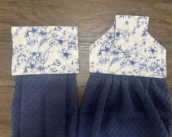 May include: Two hanging kitchen towels featuring a dark navy blue waffle-textured fabric. The top sections are made of a cream-colored fabric with a blue floral print. One towel has a folded cuff, while the other has a tab with a white button.