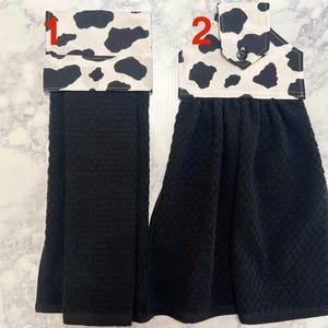 May include: Two black kitchen hand towels with a cow print top. One towel has a simple fold-over design, while the other has a button closure. The towels are made of a textured fabric.