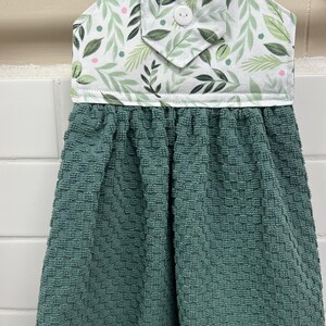 May include: A hanging kitchen towel featuring a solid dark teal, textured, waffle-weave fabric attached to a white fabric topper. The topper displays a pattern of green leaves and small pink dots, secured with a single white button at the center.