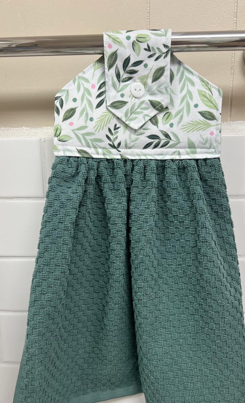 May include: A hanging kitchen towel featuring a solid dark teal, textured, waffle-weave fabric attached to a white fabric topper. The topper displays a pattern of green leaves and small pink dots, secured with a single white button at the center.