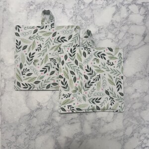 May include: Two square pot holders with loop handles. The pot holders feature a white background with a pattern of green and gray leaves and small pink dots. The edges are trimmed with a light green fabric.