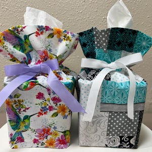 May include: Two fabric tissue box covers. The left cover features a white background with colorful hummingbirds and floral patterns, tied with a lavender ribbon. The right cover displays a teal, black, white, and gray patchwork design with a white ribbon.
